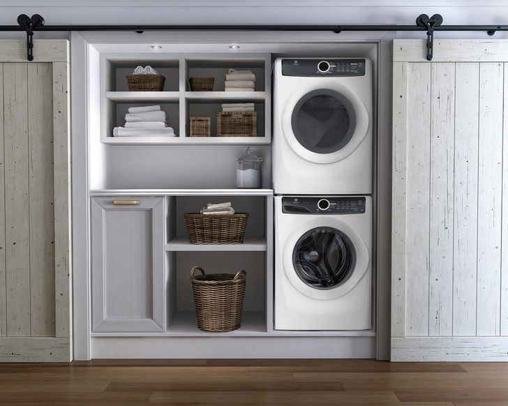 Hallway laundry room with stacked inset white washer and dryer, open upper shelving, a folding counter, lower grey cabinets, and barn-door style sliders
