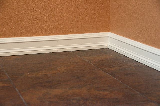 A closeup of white baseboard running along an orange wall and brown tiled flooring
