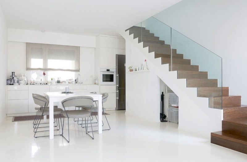White painted kitchen, white table, lower white kitchen cupboards with window above, and a brown-step staircase on the right with glass railing