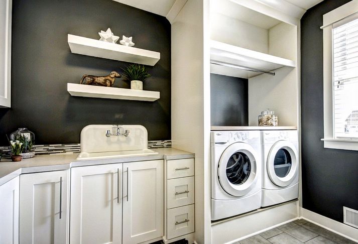 Black painted laundry room with lower white cabinets and floating shelves on the left, and a white washer and dryer on the right with a rod and shelf above