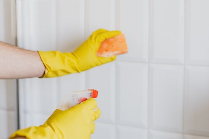 Gloved hands holding a sponge and spray bottle to clean ceramic tile walls