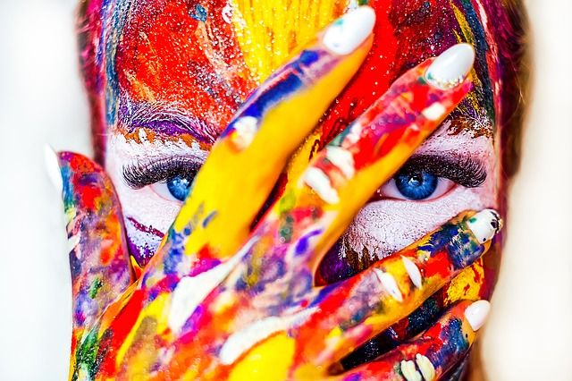 A woman with blue eyes who has her right hand partially over her face that's covered in multiple colours of paint