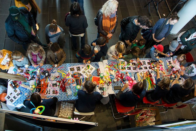 A table full of crafts materials surrounded by kids and a volunteer helping them create pulsars