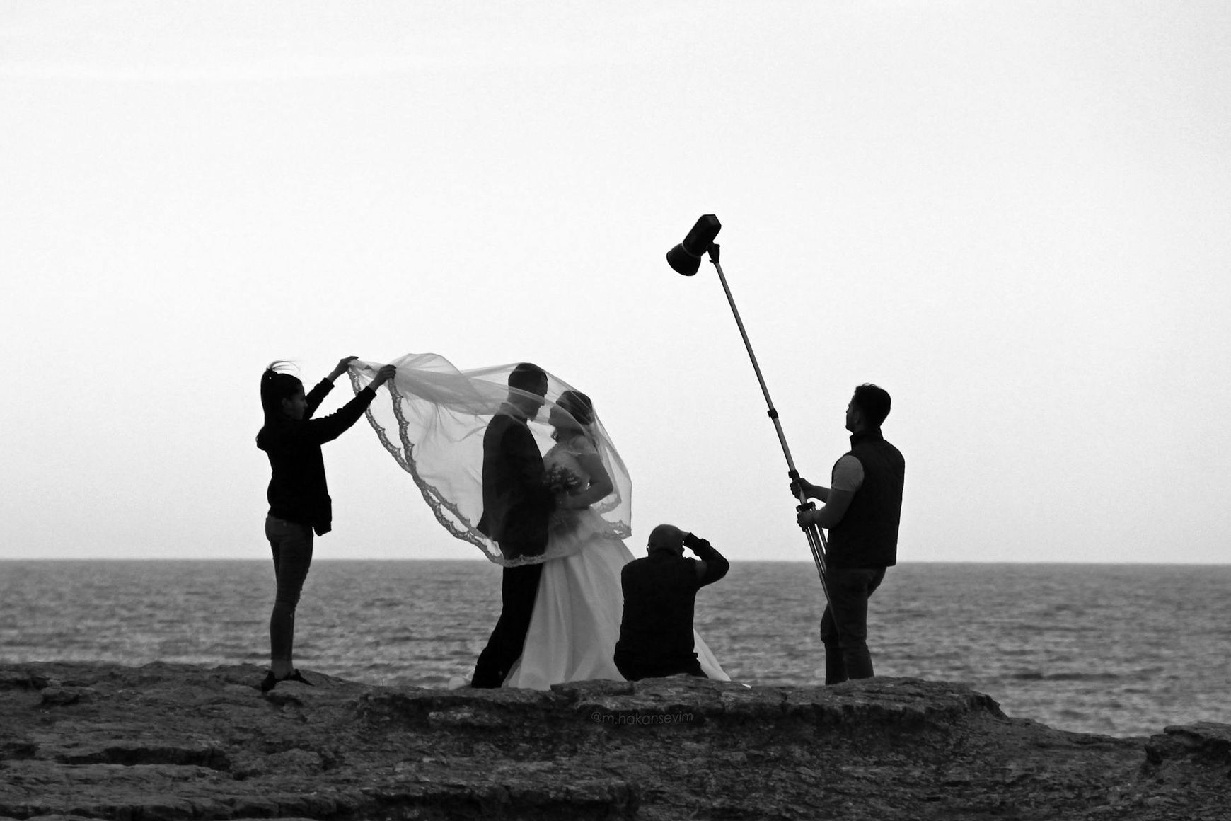 Wedding photographer image in black and white showing photographer on rocks with bride and groom and two assistants, one holding a light.