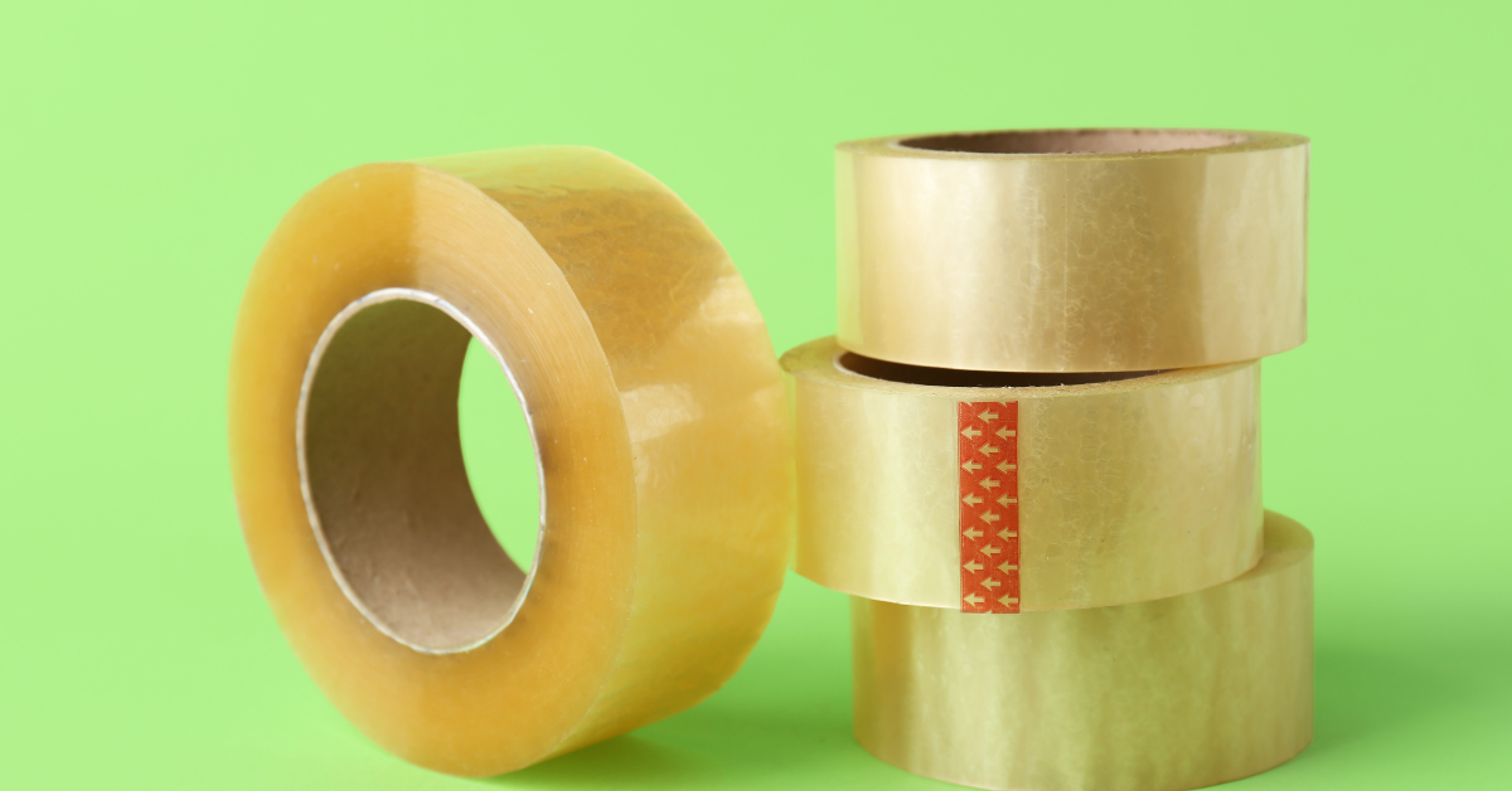 Four rolls of clear packing tape on a bright green background, one roll upright and three stacked.