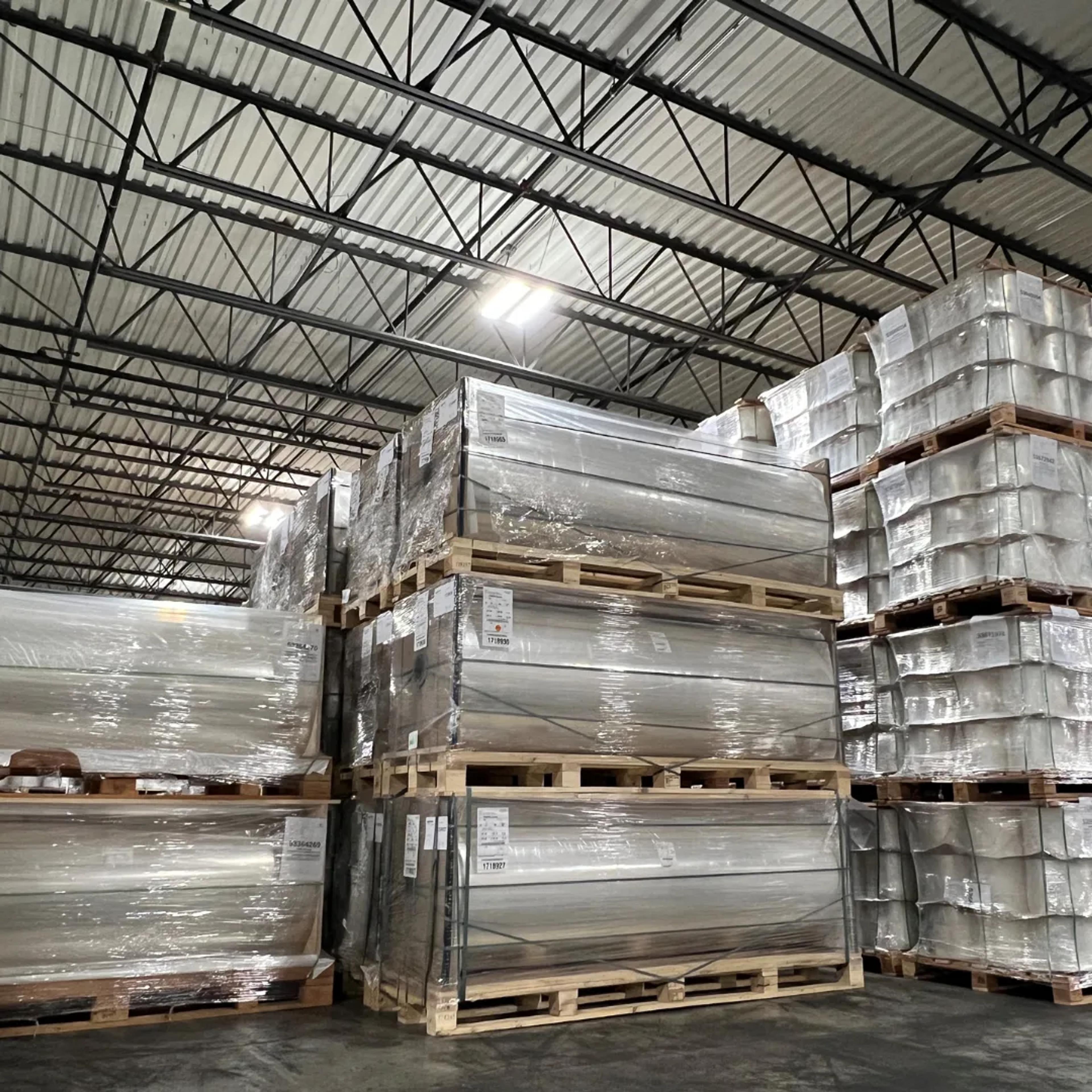 Stacked pallets of large plastic-wrapped flexible film rolls inside a high-ceiling warehouse.