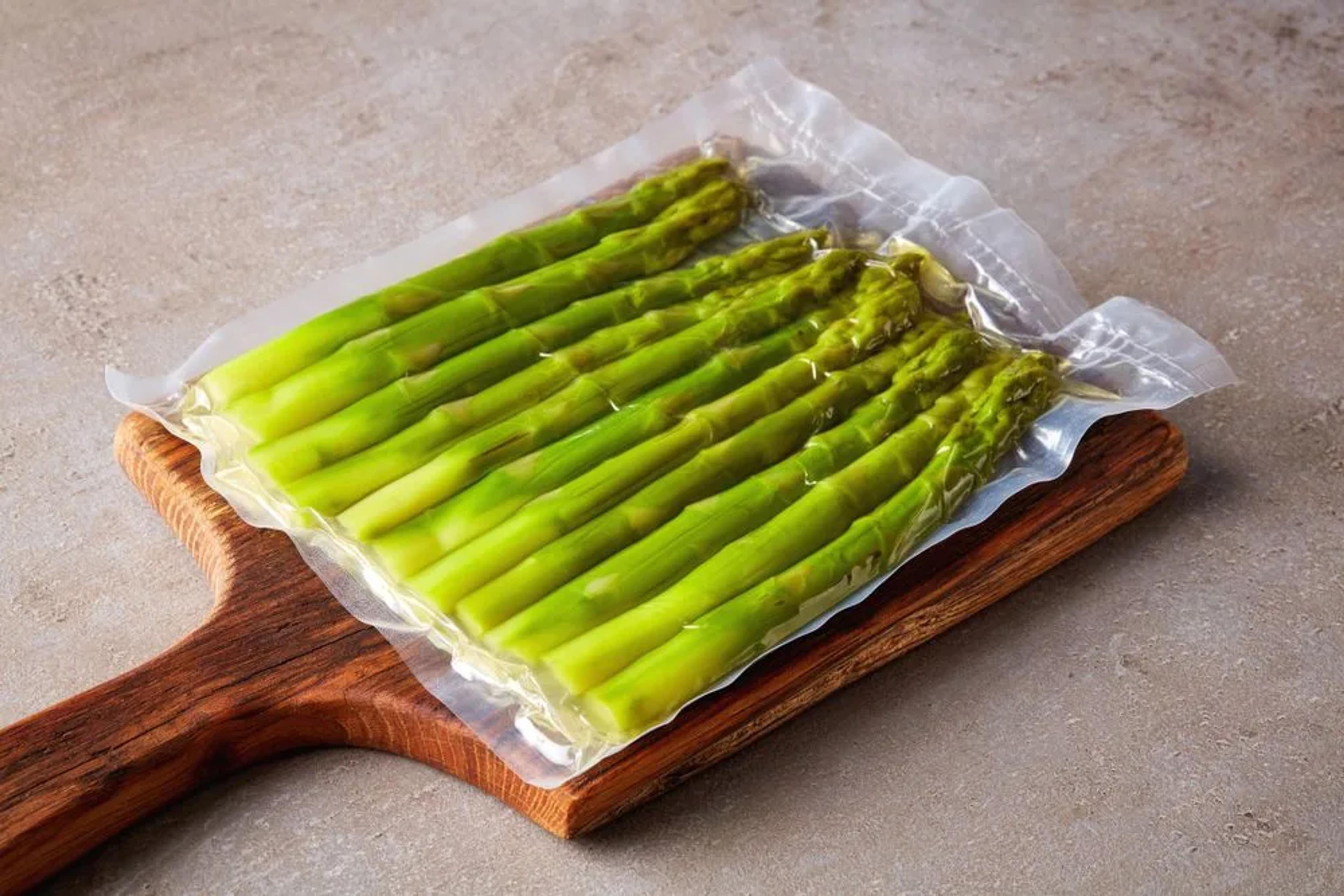 Asparagus spears vacuum-sealed in a clear flexible pouch on a wooden cutting board.