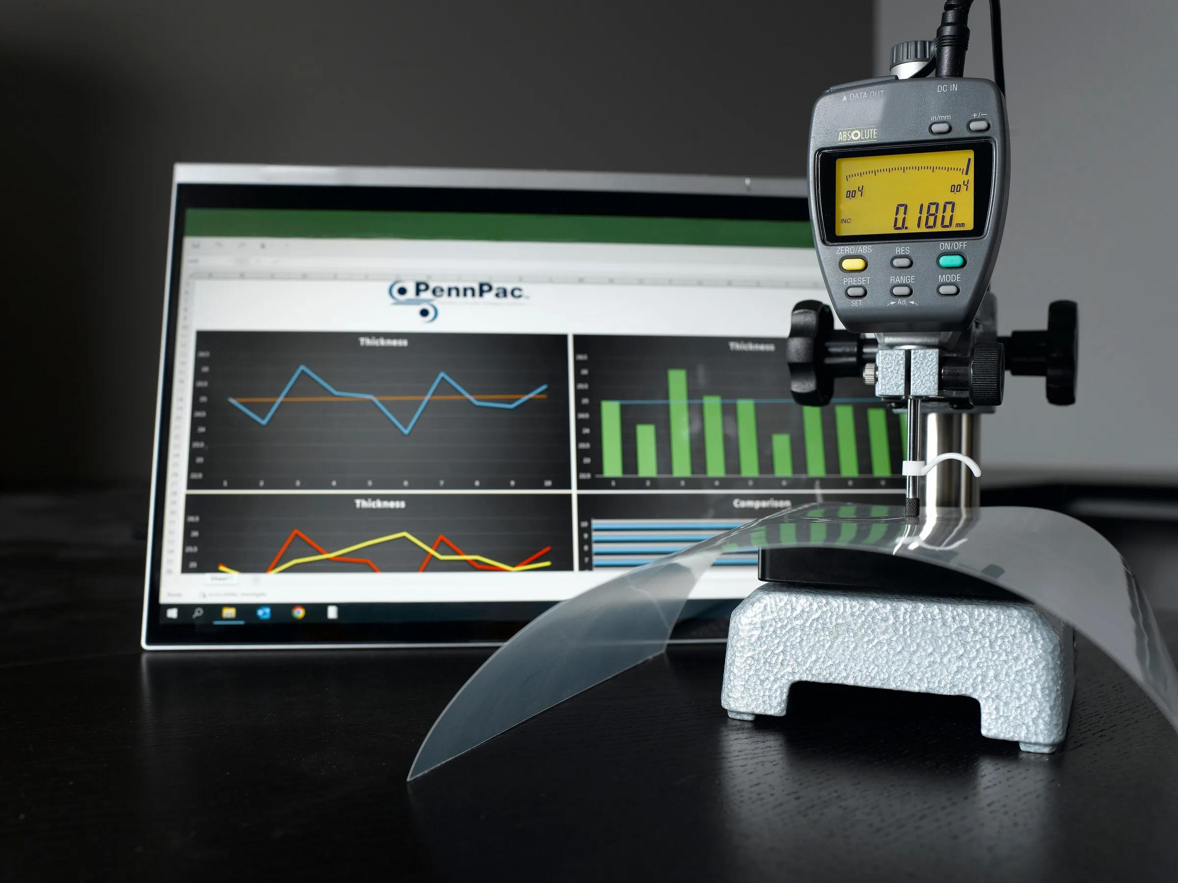 Film thickness measurement with PennPac analytics Digital micrometer measuring clear flexible film sample on a stand with a laptop showing PennPac thickness charts in the background.