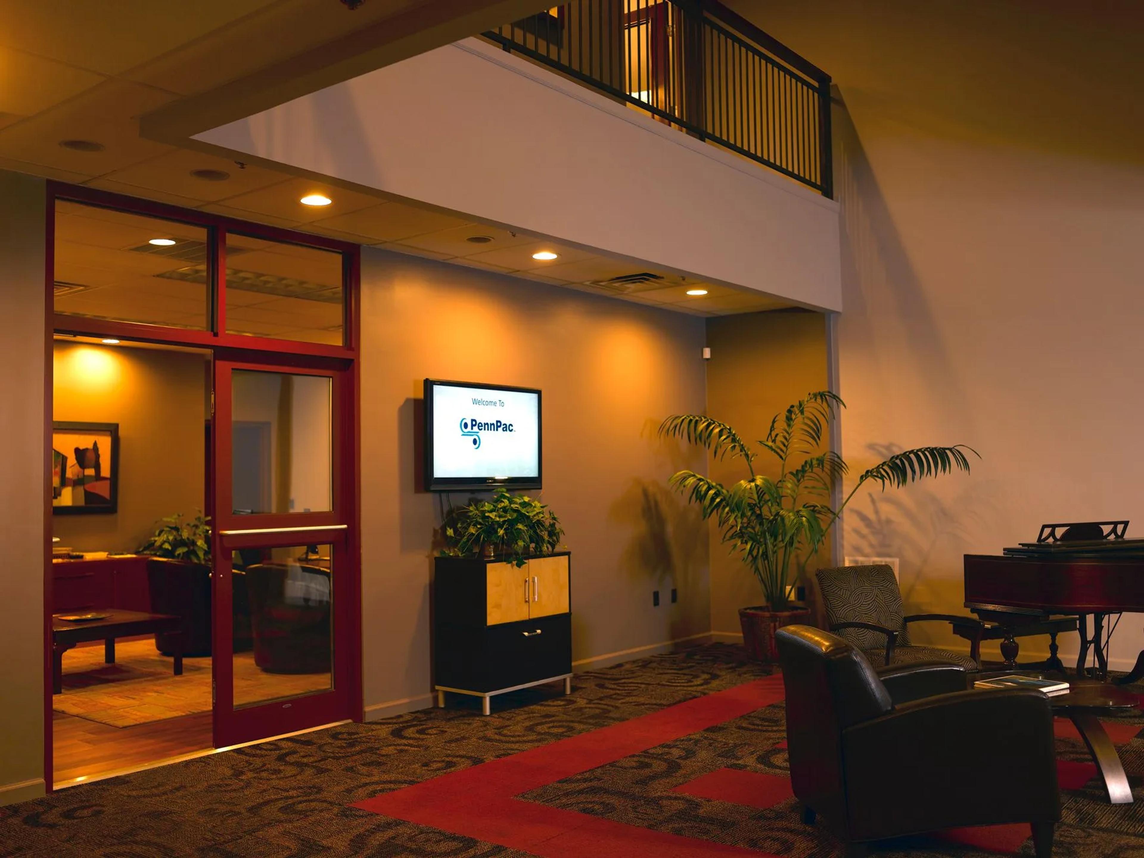 Corporate lobby with leather chairs, wall-mounted monitor reading 'Welcome to PennPac', red-framed glass doors and potted plants.