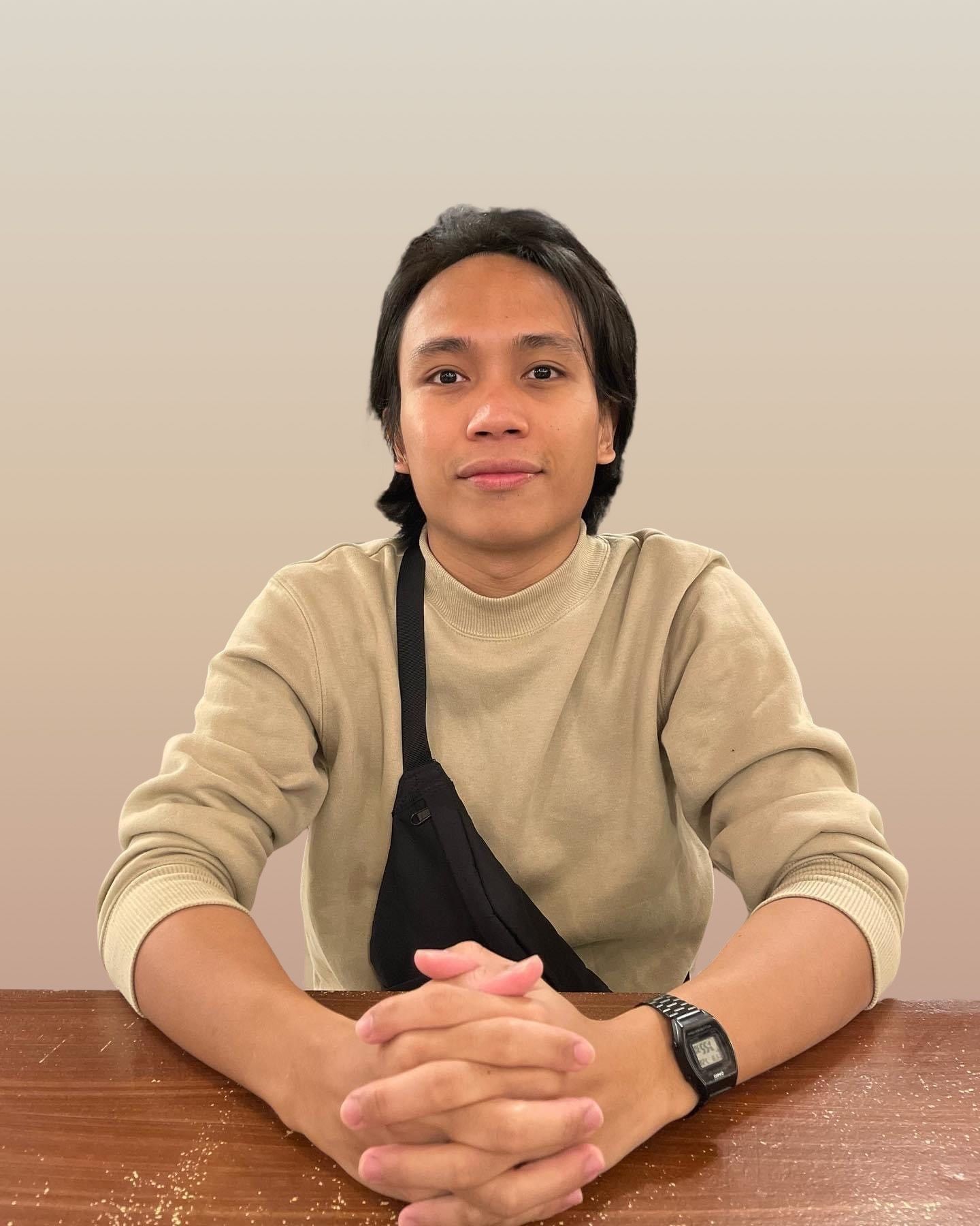 Portrait of Elloani Pitogo, wearing beige sweatshirt, seated at a table with hands clasped
