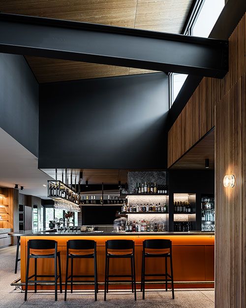 Contemporary bar interior featuring a striking burnt orange counter with black stools, polished concrete flooring, and a dramatic black overhead structure. Shelving displays bottles and glassware against a dark stone backdrop with integrated lighting. Timber ceilings and vertical slatted wall panels add warmth and texture, while high clerestory windows introduce natural light to the sophisticated space.