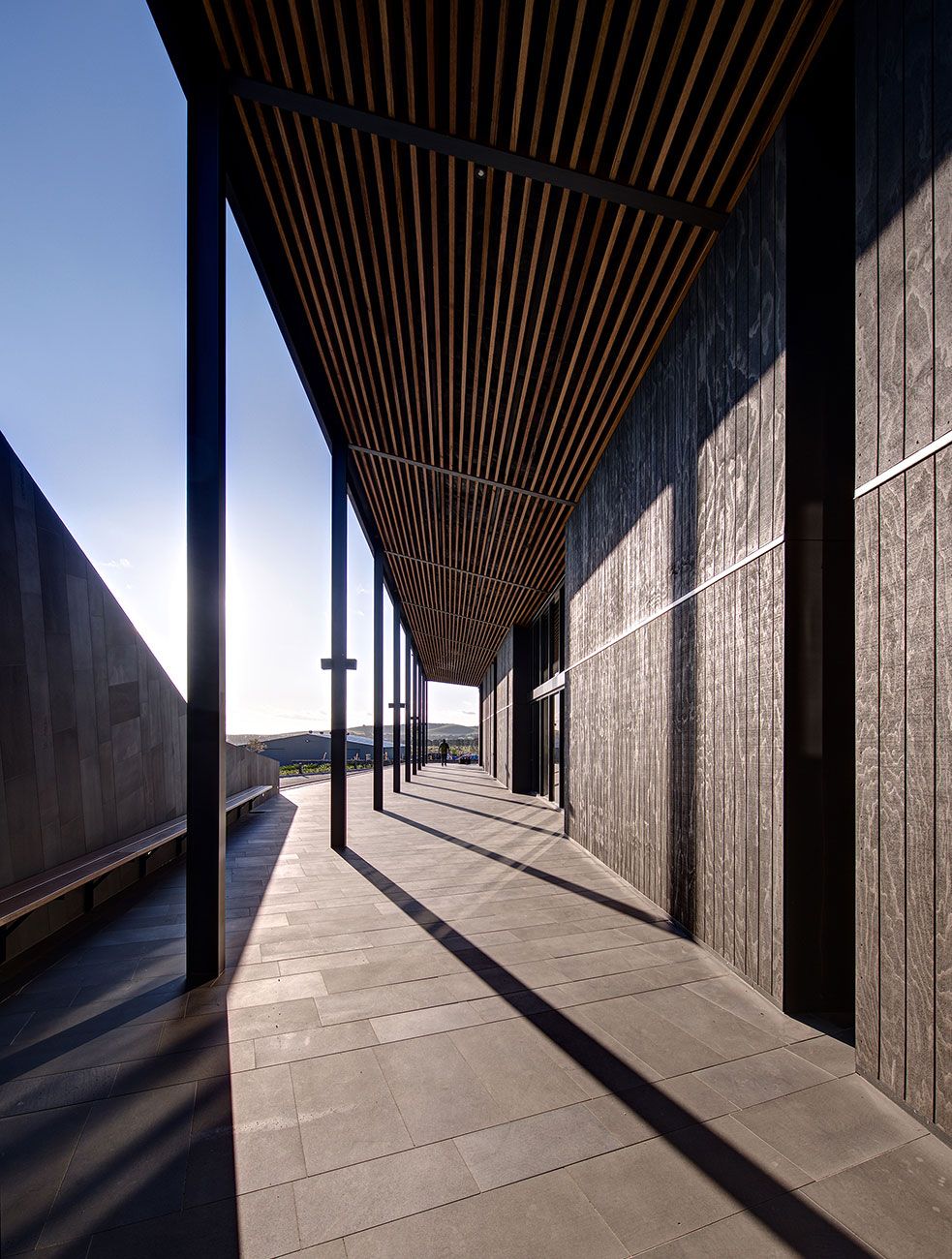 Covered outdoor walkway with vertical steel columns and a slatted timber ceiling casting linear shadows on the tiled floor, adjacent to a textured dark wall, under a clear sky.