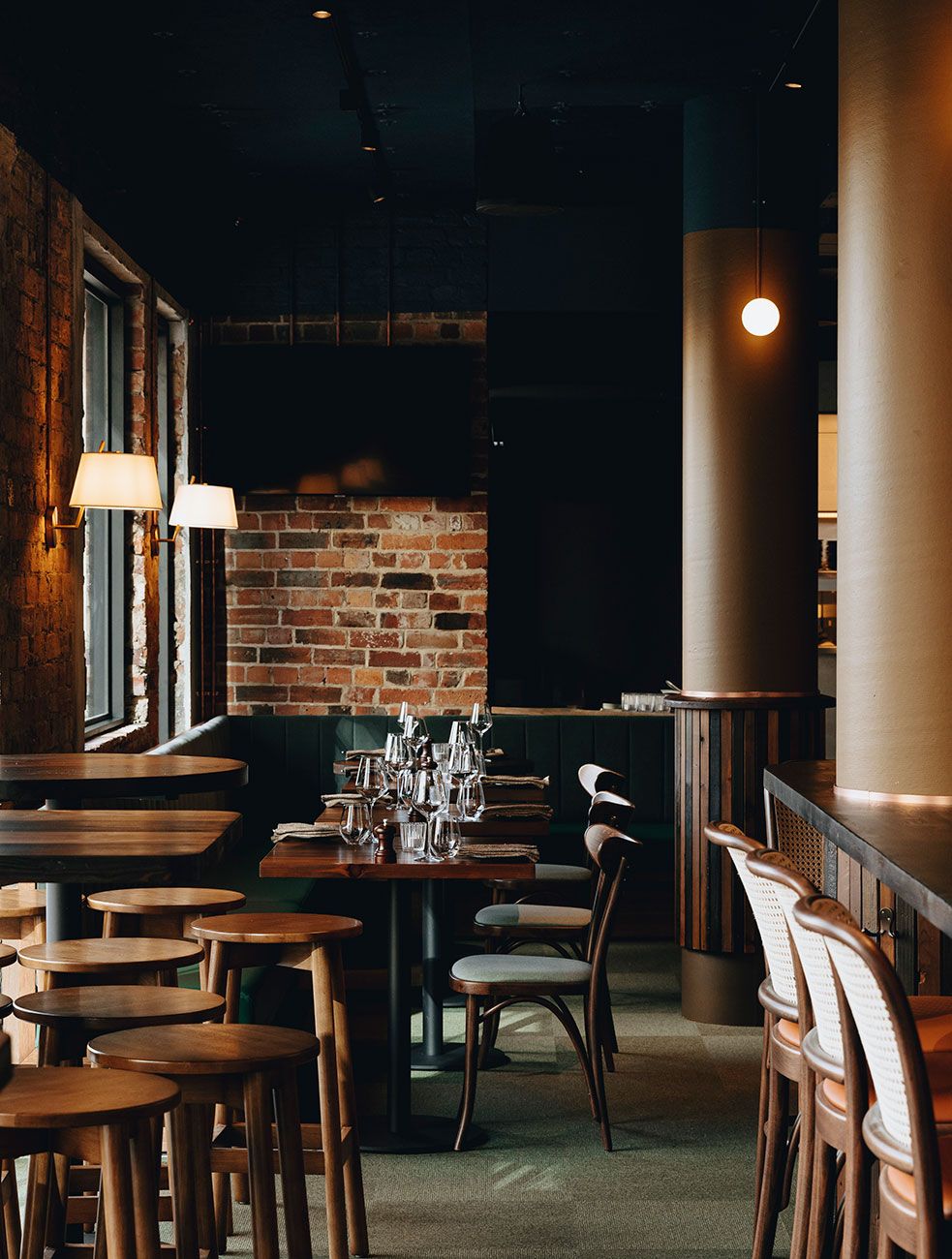 Warmly lit dining area with exposed brick walls, timber tables, and dark bentwood chairs. Bar stools with woven cane backs and tan leather seats line a curved counter with vertical timber detailing. Soft wall lamps and pendant lights add intimate illumination, while structural columns subtly define zones in this refined hospitality interior.