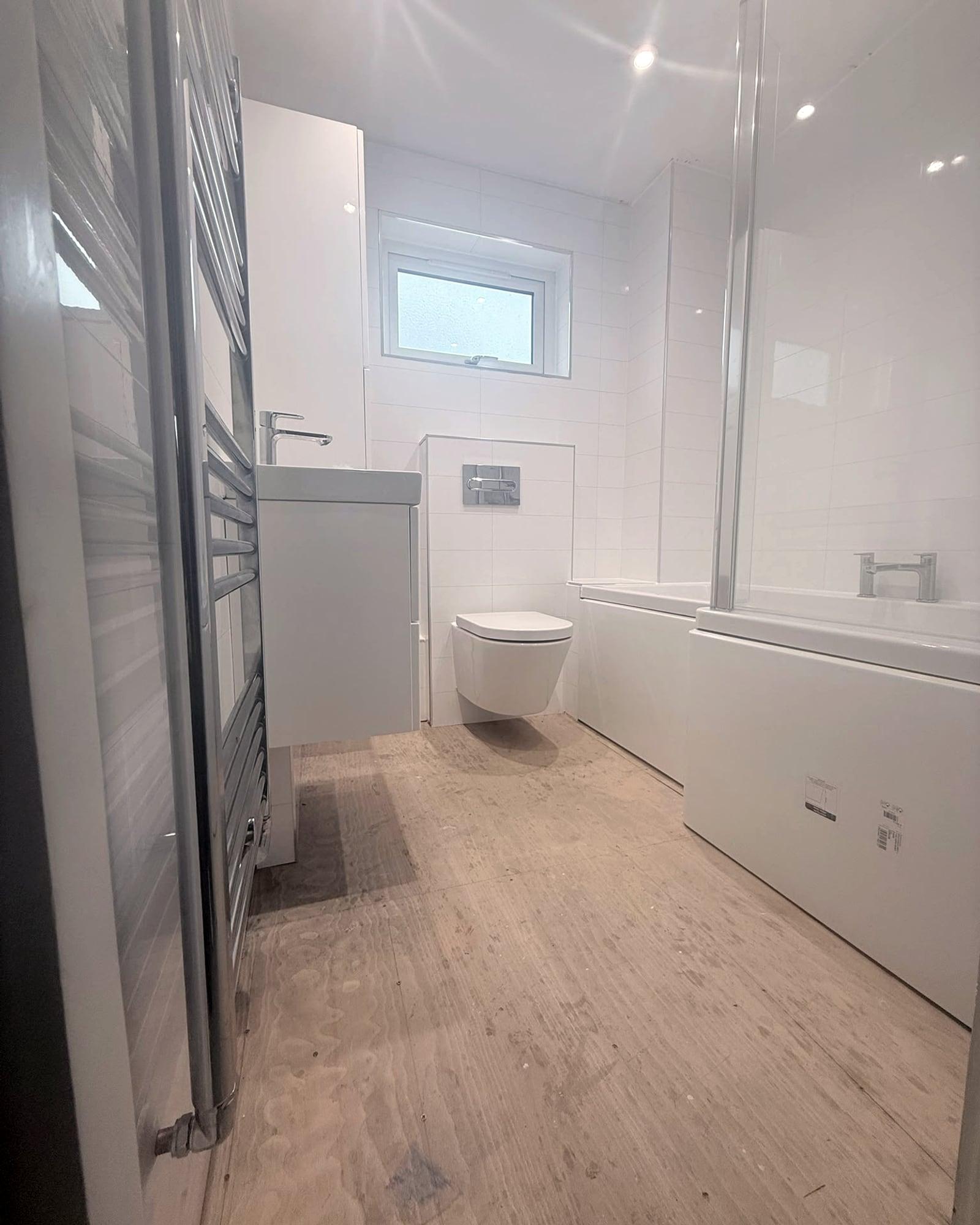 A long white bathroom: a chrome towel radiator, a basin unit, a wall-hung toilet under the window and a bath with a glass screen