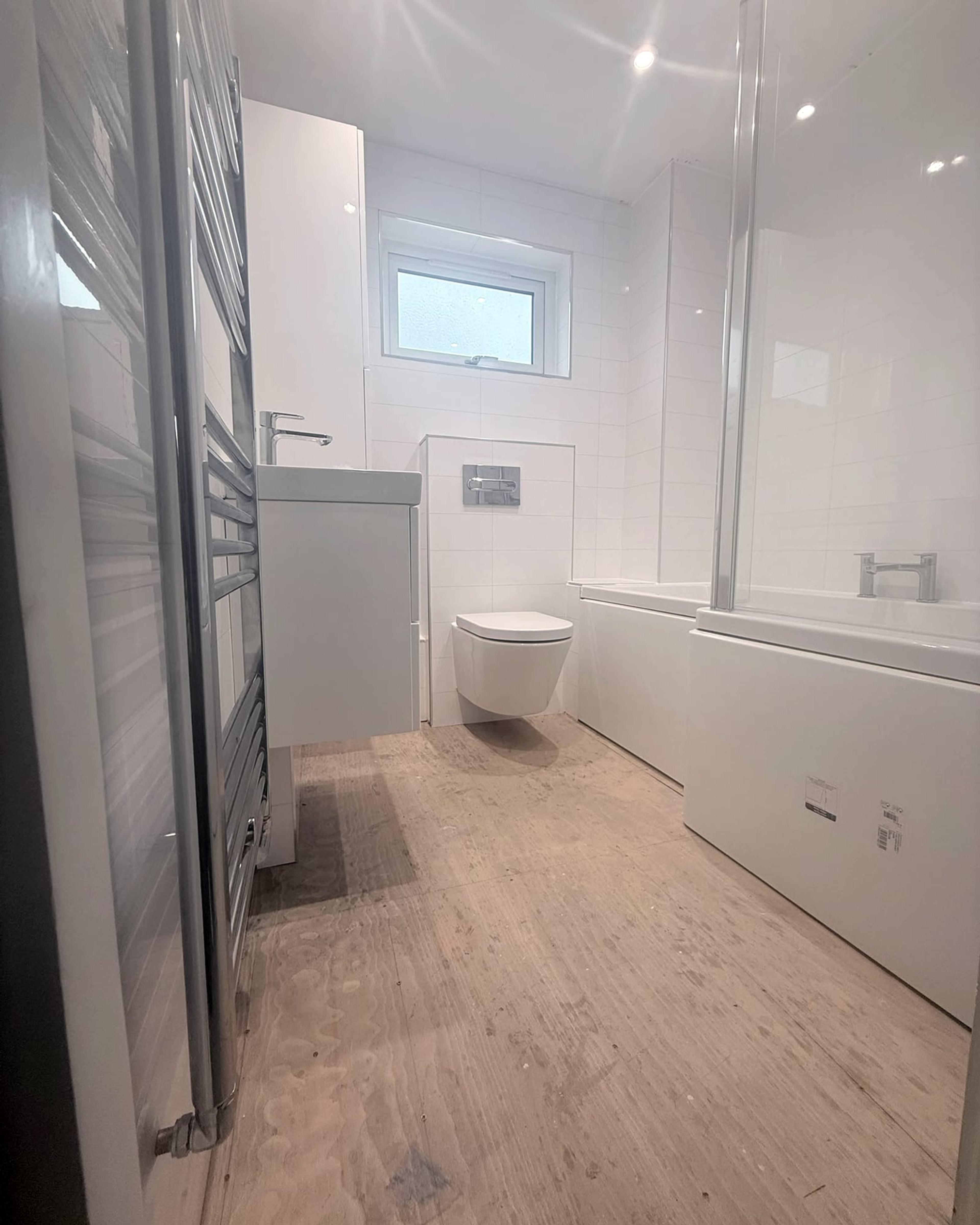A long white bathroom: a chrome towel radiator, a basin unit, a wall-hung toilet under the window and a bath with a glass screen