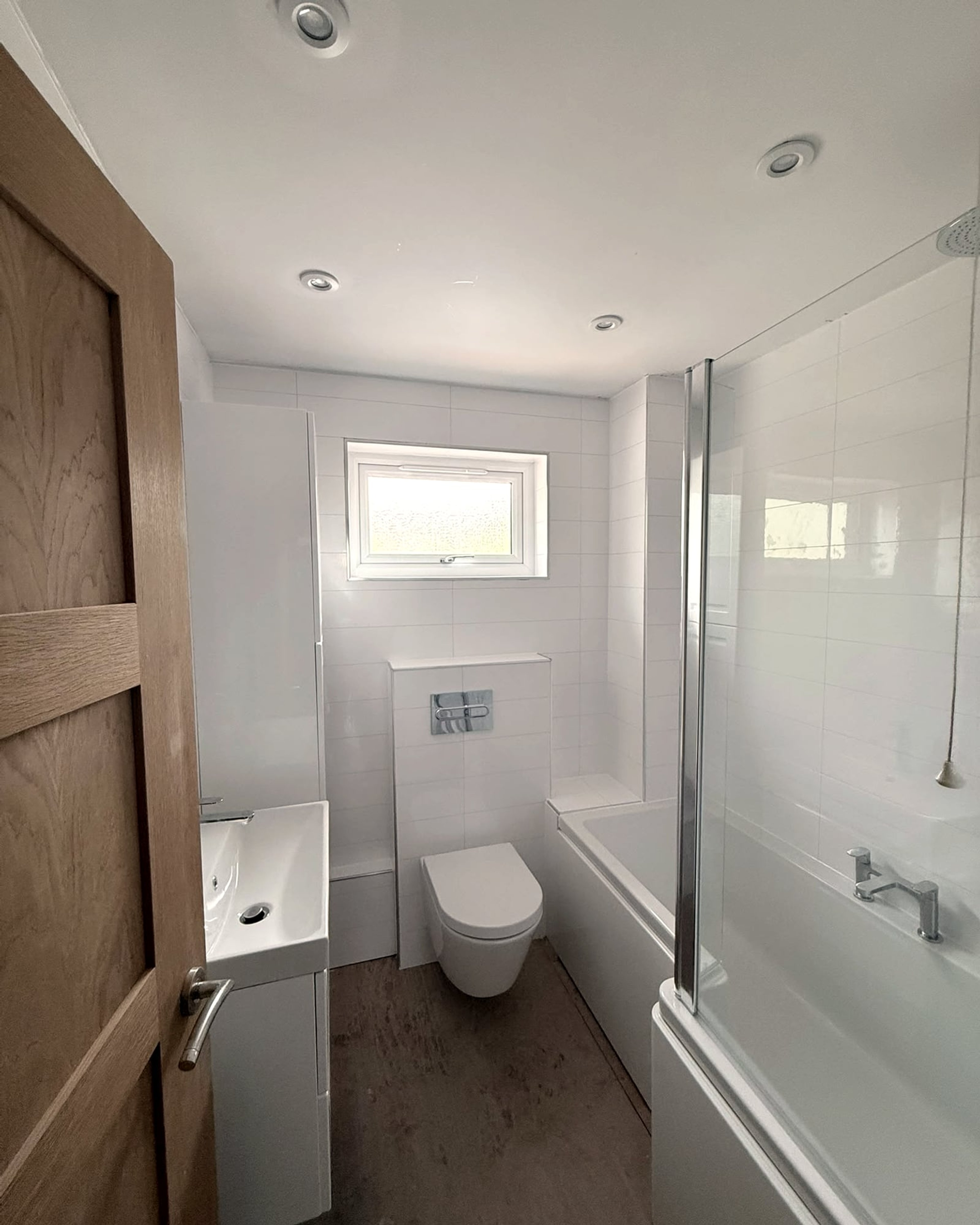 A white bathroom with a wall-hung toilet, bath with glass screen and wood-effect floor