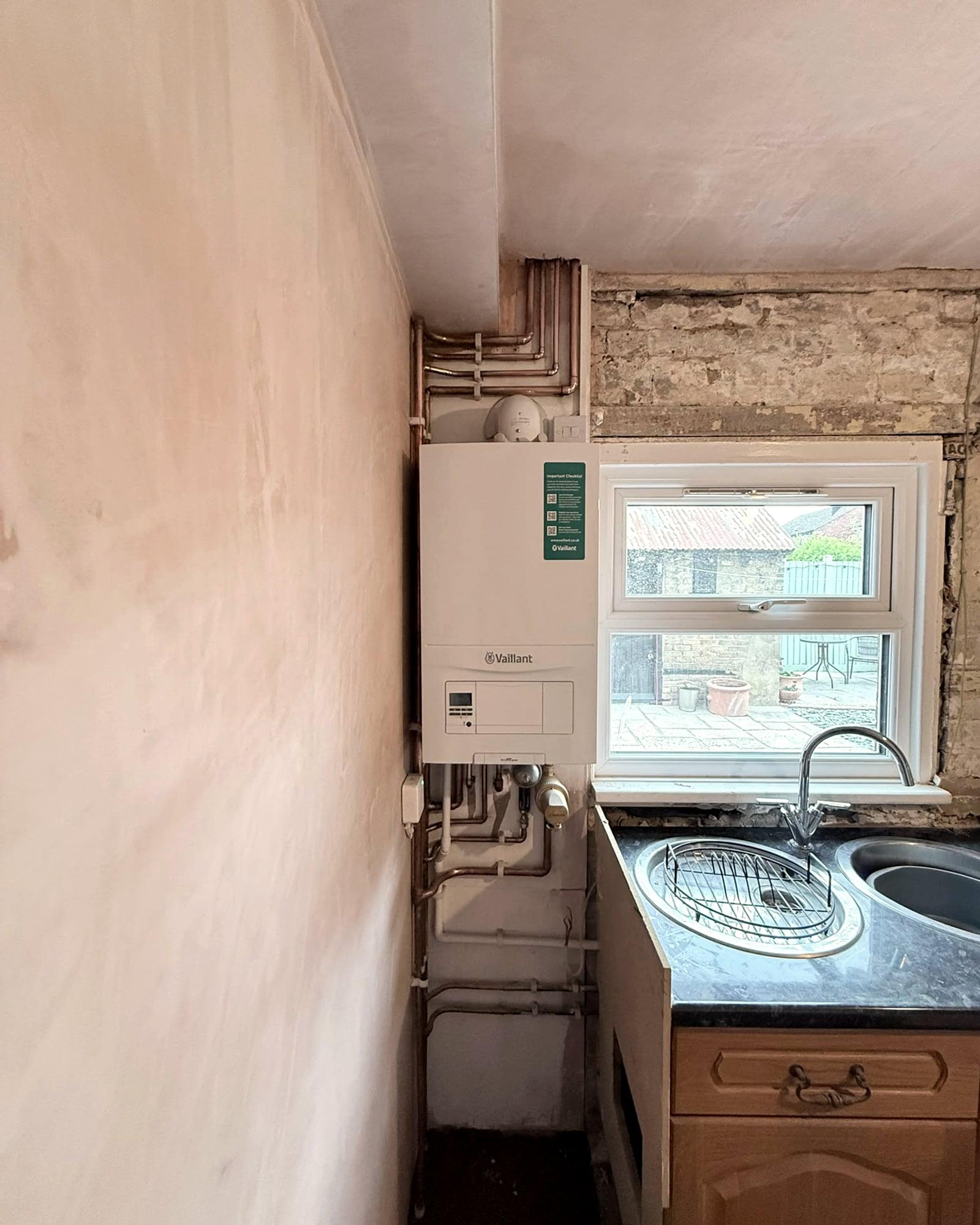 A Vaillant boiler on a freshly plastered kitchen wall beside a window, with copper pipework running into the ceiling