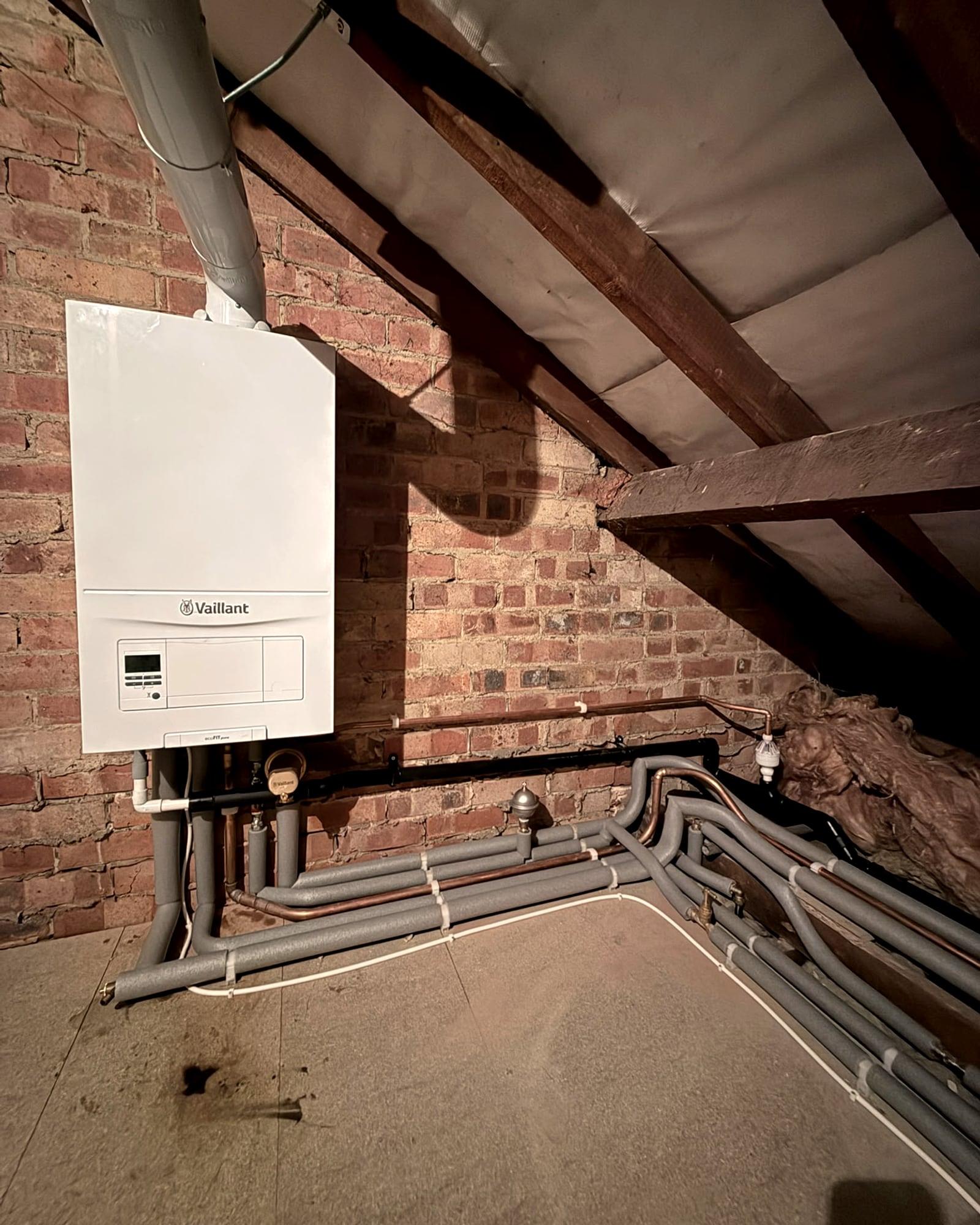 A Vaillant boiler on the brick wall of a loft under the roof timbers, with grey pipework running along the floor