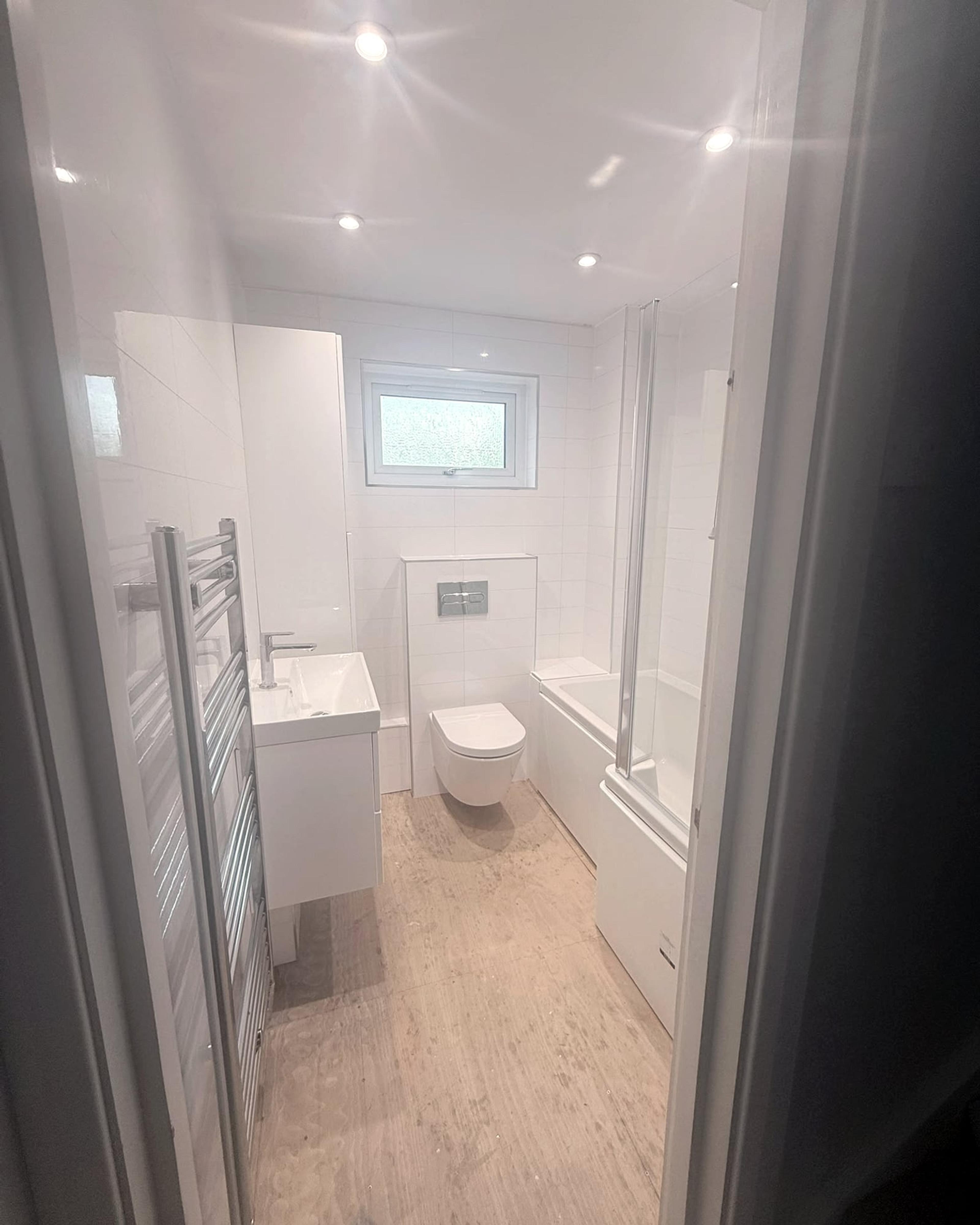 The finished bathroom seen through the doorway: white tiles, a wall-hung toilet and basin, and the bath along the wall