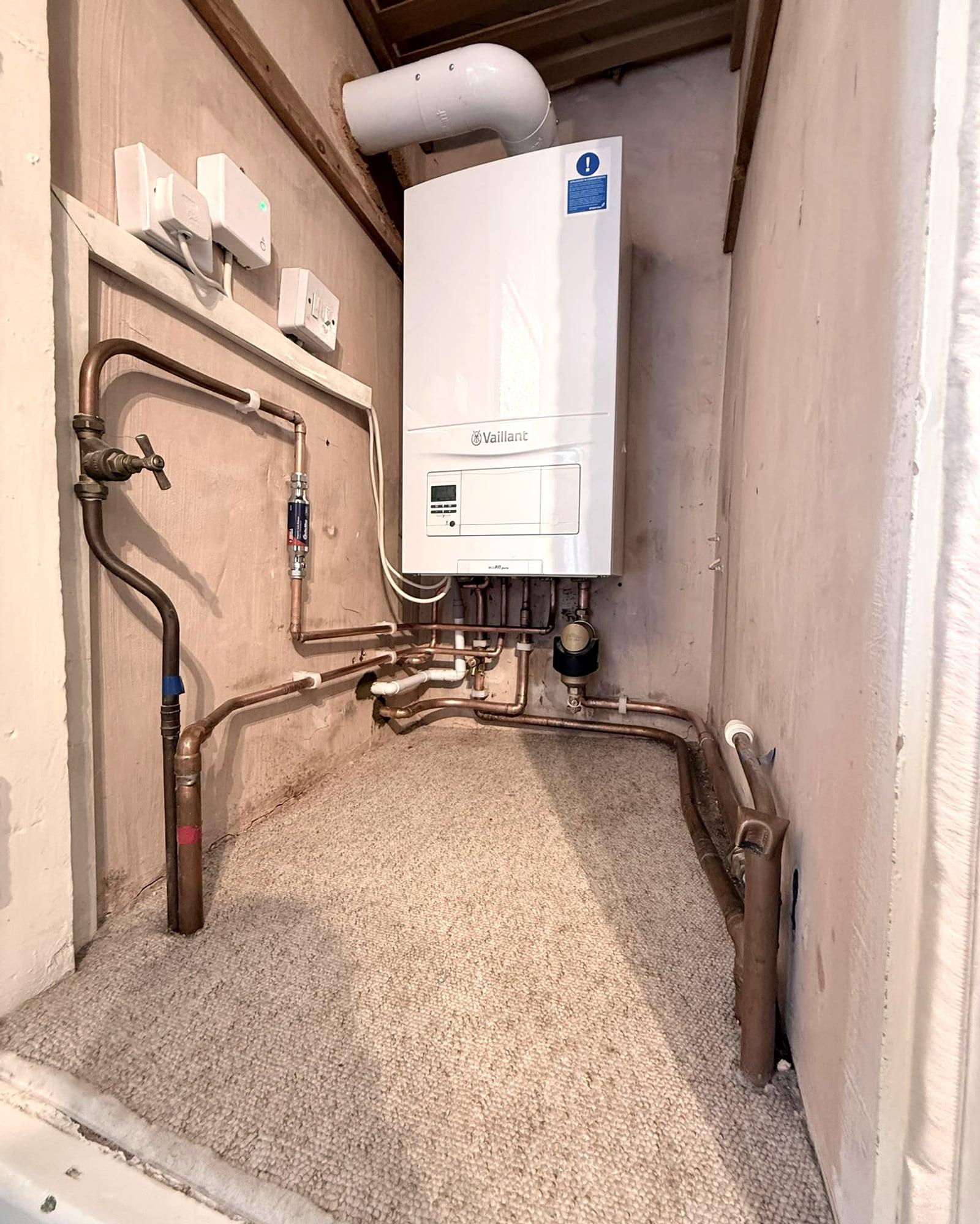 A Vaillant combi boiler in a cupboard with bent copper pipework sweeping across a carpeted shelf