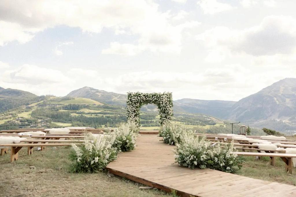 Outdoor wedding ceremony setup with a wooden aisle, white floral arch, rustic wooden benches, and a mountain landscape in the background under a partly cloudy sky.