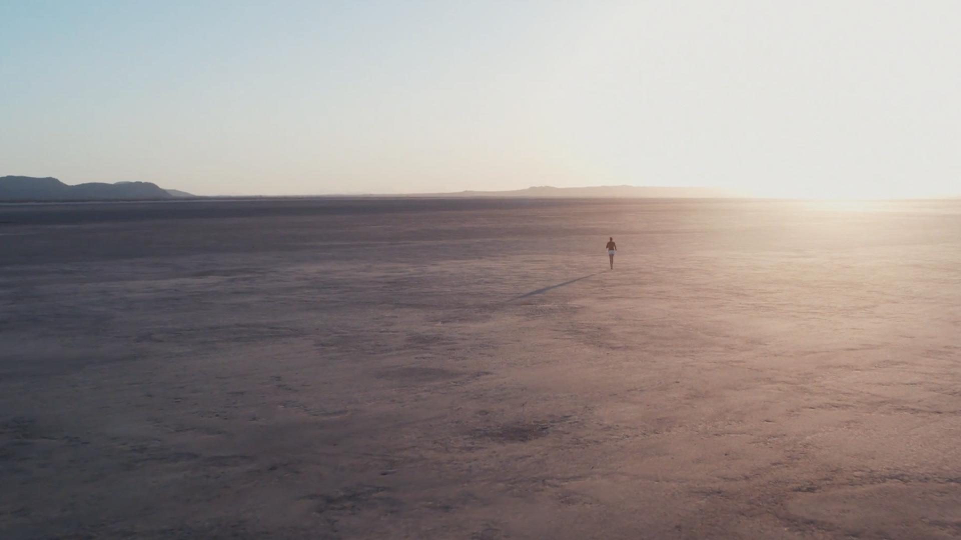Far off person in what looks to be a desert with a sun flare in the back.