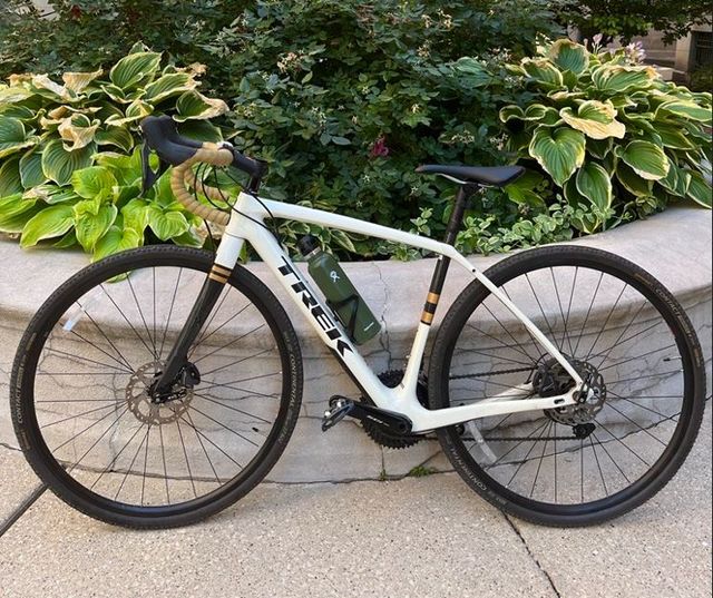 Photo of a white Trek bike with drop bars on a sidewalk, against a background of vibrant green foliage.