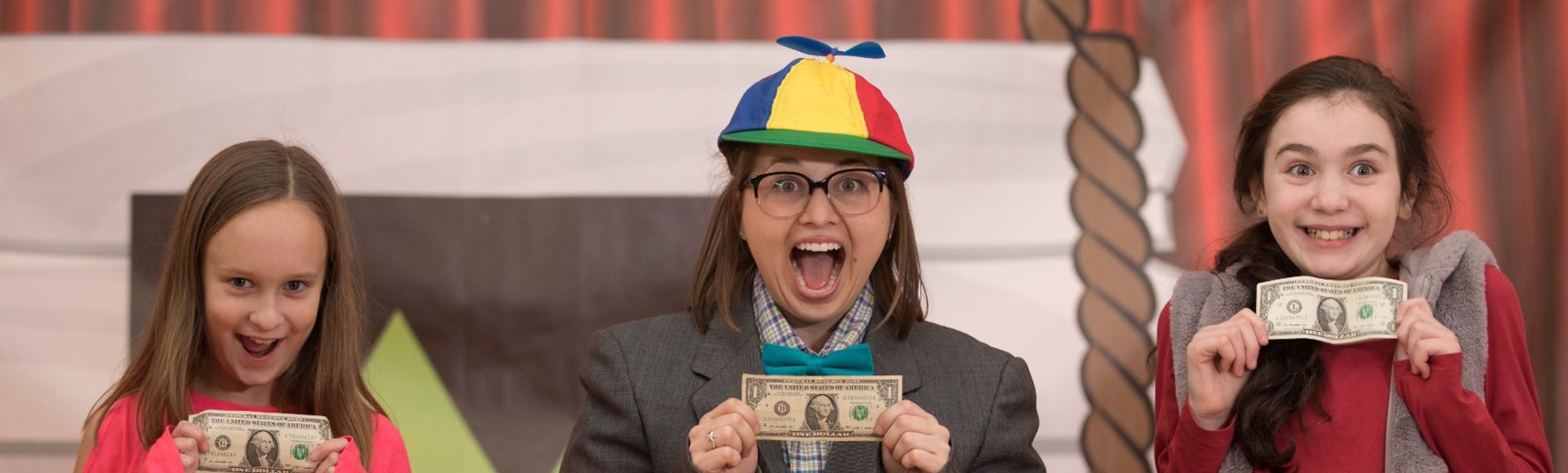 children's theater performer in a funny hat holding up dollar bills with two kids
