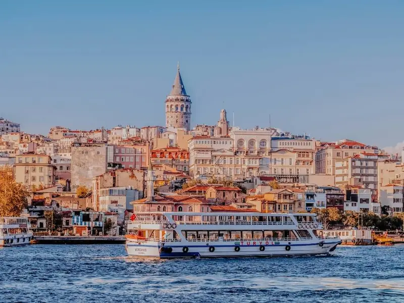 View across the Bosphorus in Istanbul with a white passenger ferry cruising over deep blue water, set against a skyline of warm-toned buildings and the Galata Tower rising above the rooftops under a clear sky. The scene captures Istanbul’s mix of waterfront energy, historic architecture, and big-city atmosphere, perfect as a first-trip wildcard for travellers chasing culture, food spots, and easy ferry rides between neighbourhoods.