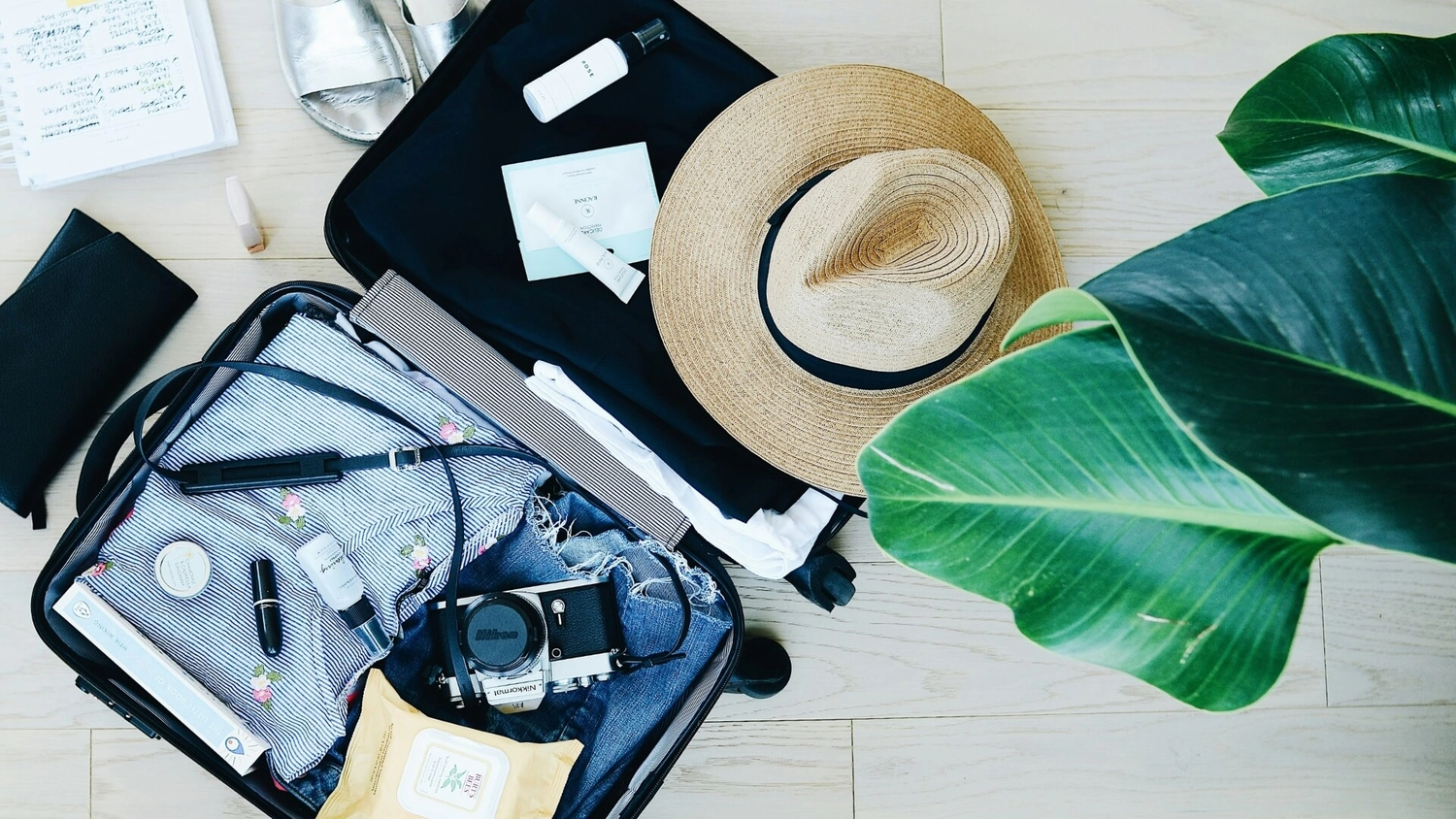 Open suitcase with premium holiday essentials including folded resort clothing, straw sun hat, camera, sandals, skincare and tropical leaves, ready for a MedSailors Premium Mediterranean sailing trip.