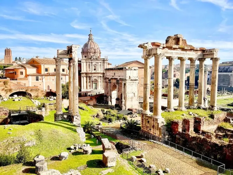 Sunlit view across the Roman Forum in Rome, with ancient stone columns and temple ruins in the foreground and historic domed buildings rising behind them under a bright blue sky. The scene highlights Rome’s iconic ancient history and open-air archaeological sites, perfect for first-time Europe travellers looking for culture, landmarks, and easy walking routes through the city centre.