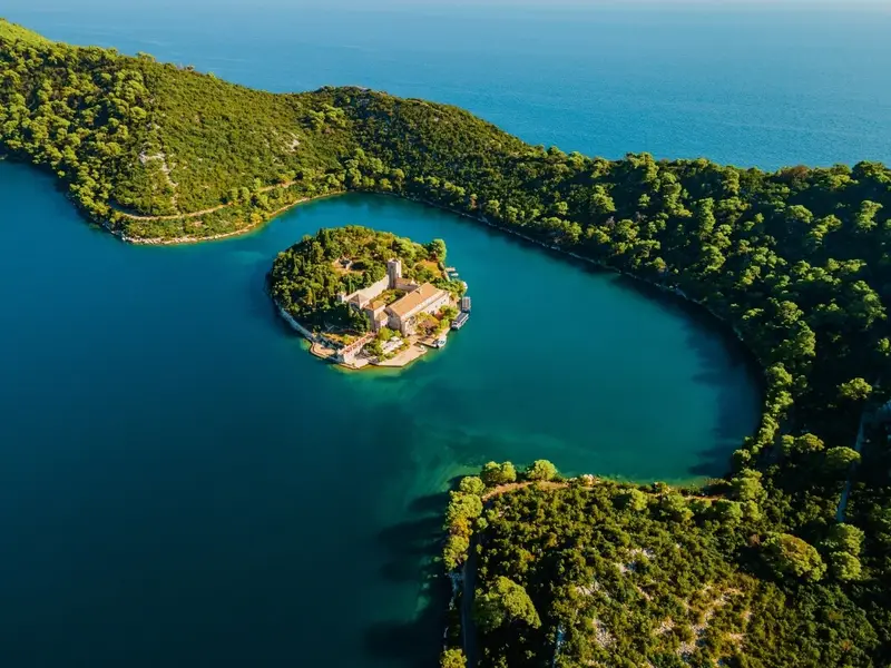 Aerial view of Mljet National Park in Croatia showing a small islet with a historic monastery surrounded by deep blue-green lakes and dense forest. Image used in a MedSailors Odyssey locations guide as a “Croatia rumoured cave” add-on, referencing local tradition that links Mljet to Calypso and Odysseus, and highlighting how the wider Adriatic and Ionian region is full of real landscapes that match the sea-journey vibe of Homer’s The Odyssey.