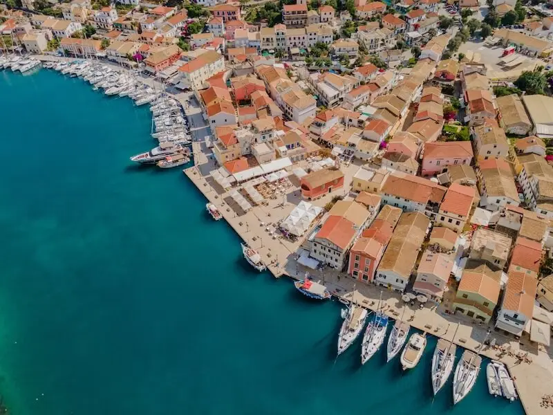 Aerial view of Gaios on Paxos Island, Greece, showing colourful waterfront buildings with terracotta roofs, lined cafes, and sailboats moored along the turquoise harbour — a picturesque stop on MedSailors’ Ionian sailing holidays.