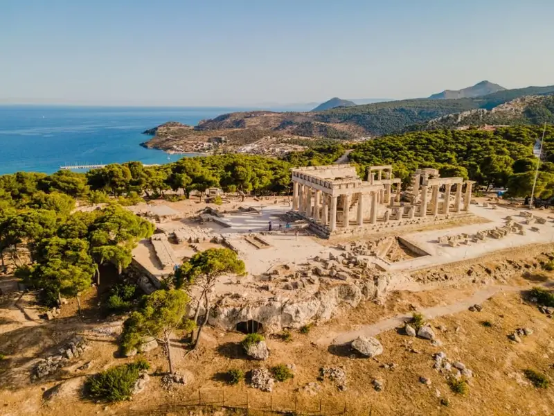 Aerial view of the Temple of Aphaia on Aegina island, Greece, showing the ancient stone columns and ruins surrounded by pine trees with the blue Saronic Gulf and coastline in the background. Image used in a MedSailors Odyssey locations in Greece guide, highlighting Aegina as an easy island trip from Athens for travellers interested in Greek mythology, ancient temples, and Homer’s world alongside Odyssey-inspired island hopping.