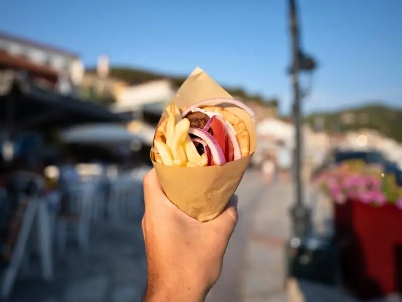 Close-up of a hand holding a freshly made Greek gyros wrapped in paper, filled with fries, onions, and pita bread, with a sunny seaside village in the background — showcasing authentic local food experiences on a MedSailors island hopping trip in Greece.