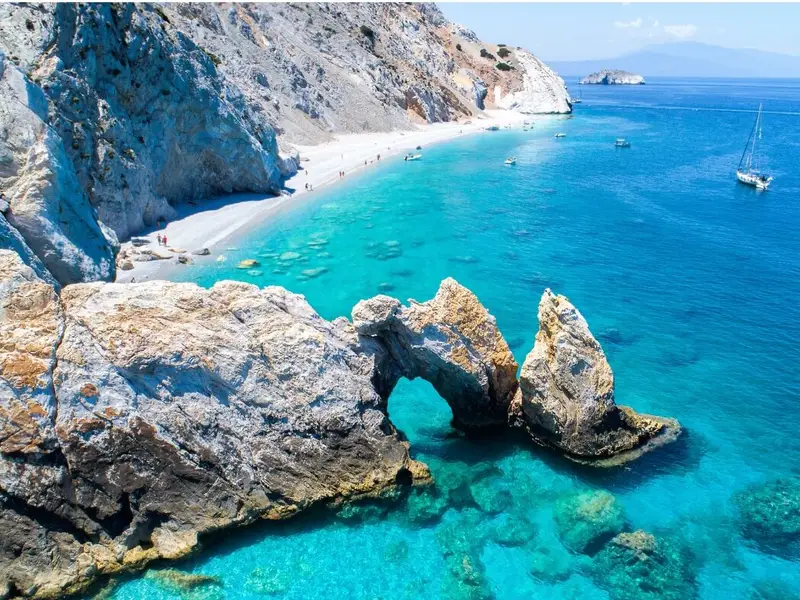 Rocky coastline and bright turquoise bay on Ithaca (Ithaki), Greece, with a small natural sea arch in the foreground, a white-pebble beach curving along the shore, and sailboats anchored in clear blue water. Image for a MedSailors guide to Odyssey locations in Greece, highlighting Ithaca as Odysseus’ home island in Homer’s The Odyssey and a must-visit stop for an Odyssey-inspired Greek island itinerary.