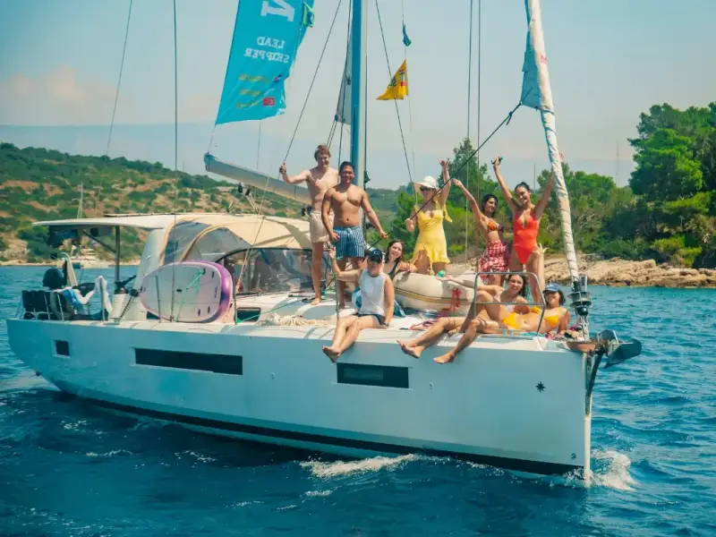 Group of young travellers celebrating on a sailing yacht during a Greek island hopping trip from Athens. Friends enjoying sunshine, turquoise Mediterranean water, and social summer vibes on an affordable sailing holiday in Greece.