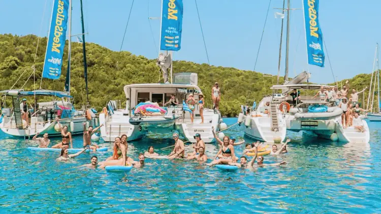 Aerial view of MedSailors travellers swimming and relaxing on colourful inflatable floats in crystal-clear Mediterranean waters during a group sailing holiday, showcasing the fun and social atmosphere of island hopping with friends.
