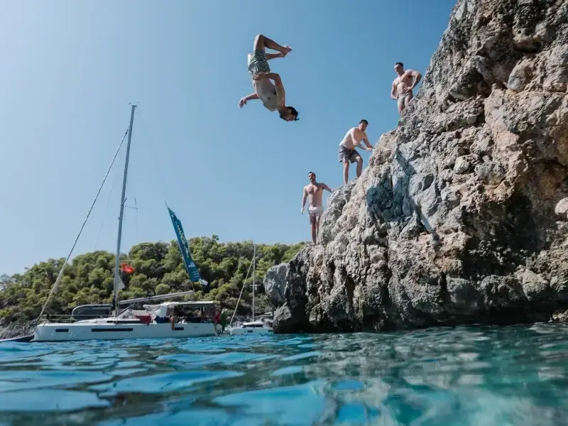 Young travellers cliff jumping into clear blue water during a sailing trip in Croatia, with a catamaran anchored nearby, capturing the adventurous side of island-hopping in Europe. This image reflects popular activities like swimming, cliff jumping, and boat days that are common on Croatia and Greece trips, where having travel insurance that covers adventure activities can be important. Ideal for illustrating Europe backpacking, sailing holidays, and active travel experiences.