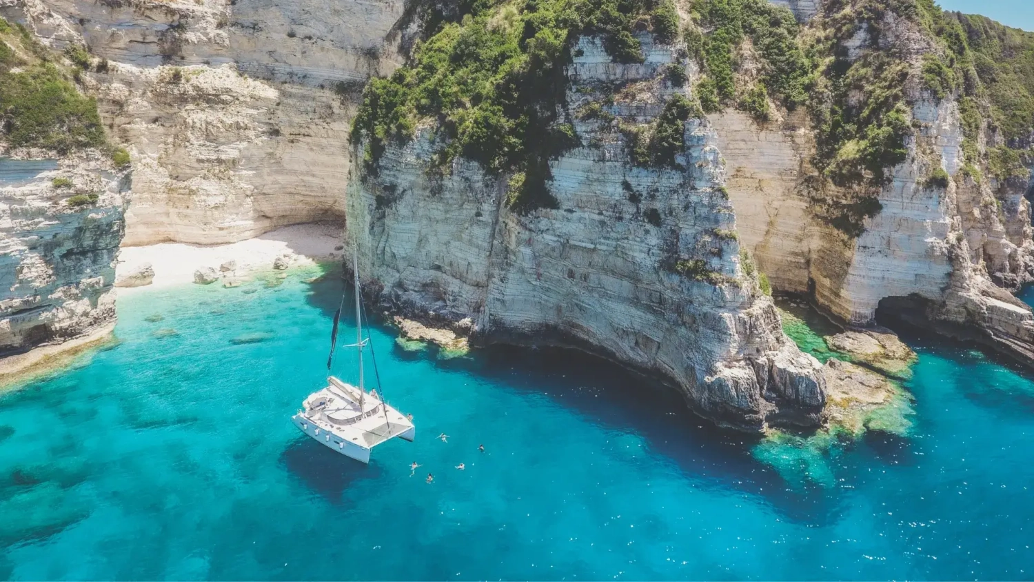 Aerial view of a sailing catamaran anchored in a bright turquoise bay beneath tall white limestone cliffs, with swimmers in the clear water near a small beach and rocky cove. Header image for a MedSailors guide to Odyssey locations in Greece, inspired by Homer’s The Odyssey and the 2026 Odyssey movie, covering real places you can visit like Ithaca, Corfu, Athens and the Saronic Islands.