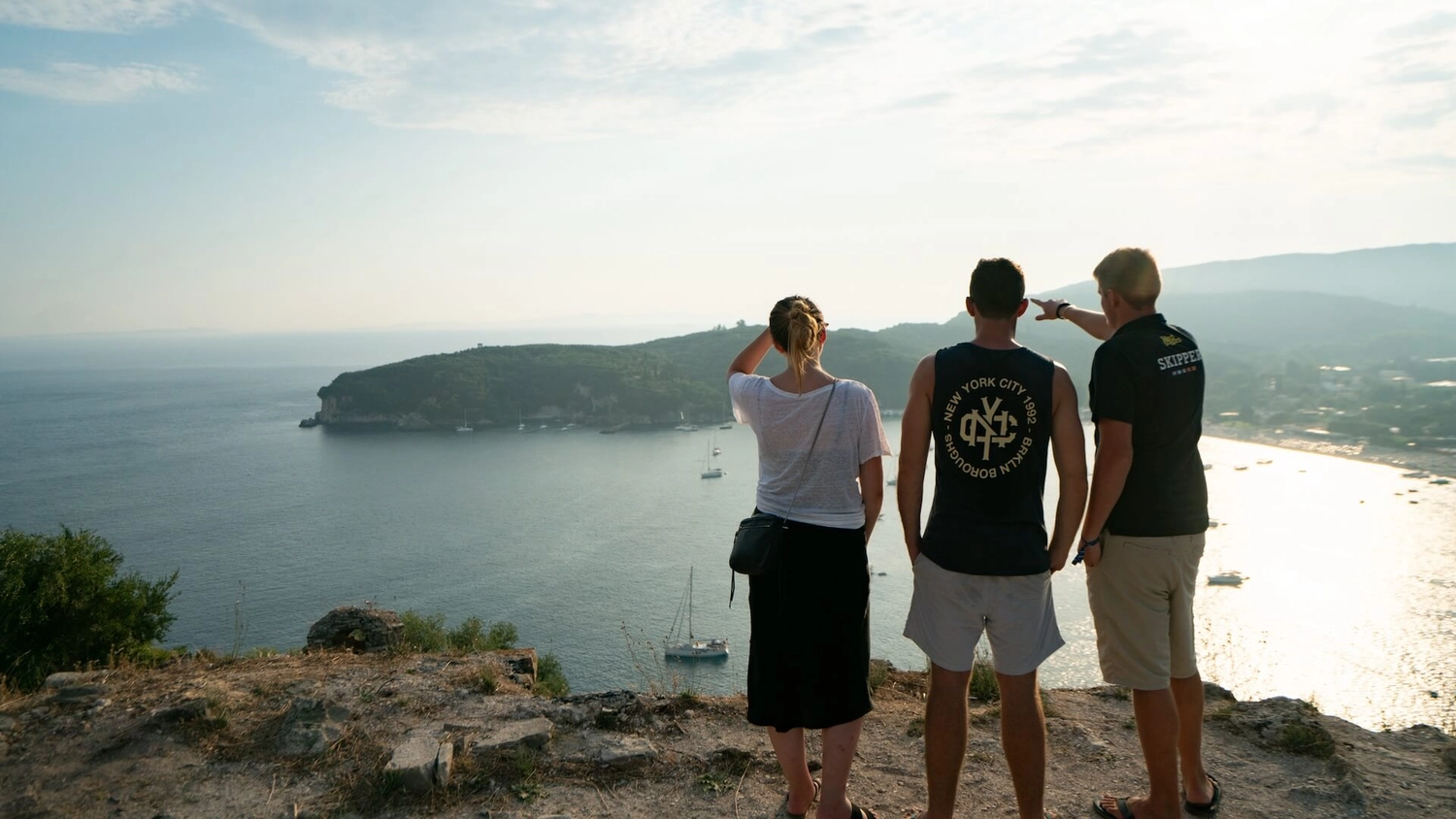 Three people standing on a cliff edge watching sunset over a bay from Parga Fortress in Greece.