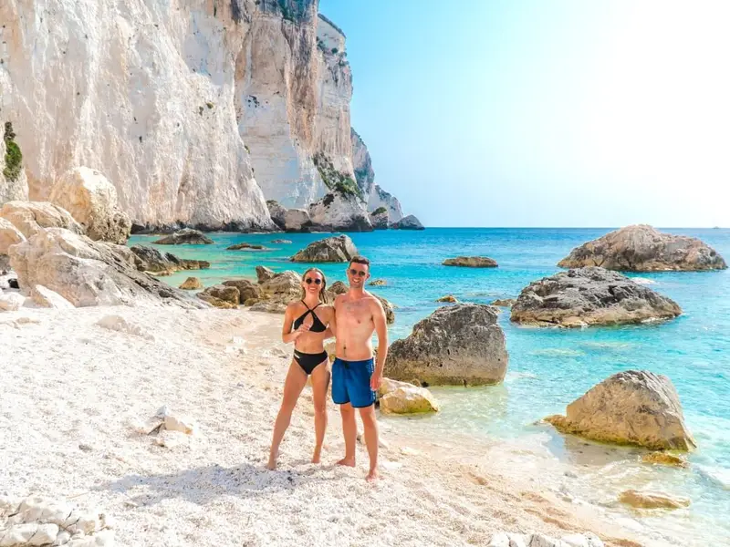 Couple standing on a bright white-pebble beach in Kefalonia, Greece, with dramatic pale cliffs, large shoreline rocks and clear turquoise Ionian water in the background. Image used in a MedSailors Odyssey locations in Greece guide to highlight Kefalonia as a stunning Ionian island stop near Ithaca, ideal for an Odyssey-inspired itinerary with beaches, coastal swims, boat days and island hopping across the Ionian Islands.