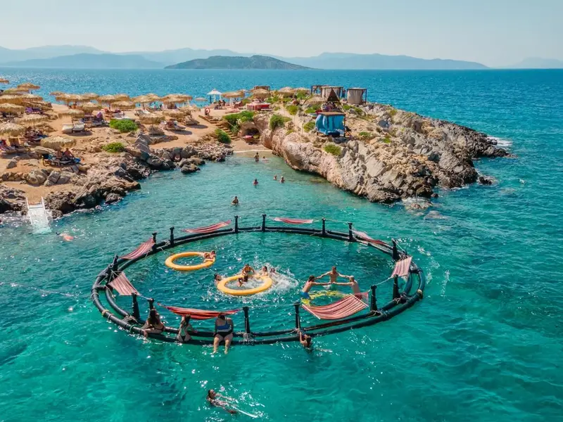 Aerial view of travellers swimming and relaxing in a circular floating sea platform in clear turquoise water off Agistri in the Saronic Gulf, with a rocky beach area, sun loungers and parasols on the shoreline behind. Image used in a MedSailors Odyssey locations in Greece guide to show the modern “island hopping near Athens” side of the trip, with easy swim stops, beach days, and group travel vibes alongside myth-inspired destinations like Aegina, Poros and Athens.
