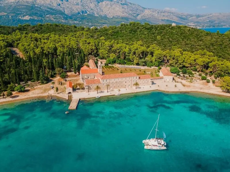 Aerial view of the Badija Monastery near Korčula in Croatia with a catamaran anchored in bright turquoise water, a scenic stop on Croatia island hopping routes.