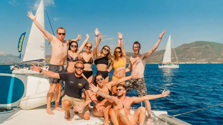Group of budget travellers on a Greek island hopping adventure, with their hands in the air smiling while on a catamaran with MedSailors.