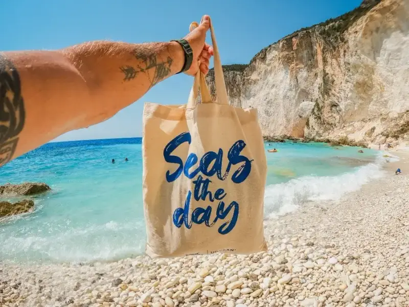 A hand holding up a MedSailors Seas the Day canvas tote bag on a stunning pebble beach in Greece with turquoise water and white limestone cliffs in the background.