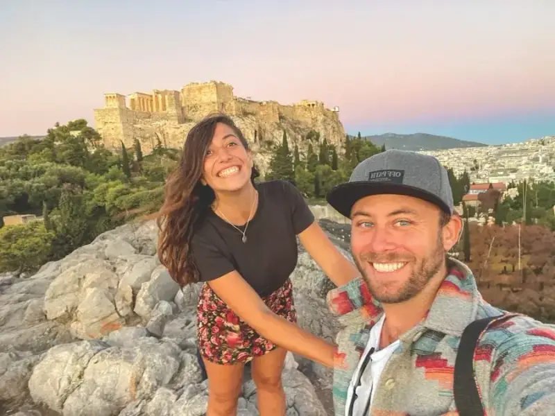Smiling couple taking a selfie at sunset with the Acropolis and the Parthenon on the hill behind them in Athens, Greece. Image for a MedSailors guide to Odyssey locations you can actually visit in Greece, using Athens as the starting point for an Odyssey-inspired itinerary before heading to islands like Aegina, Poros, Corfu and Ithaca.