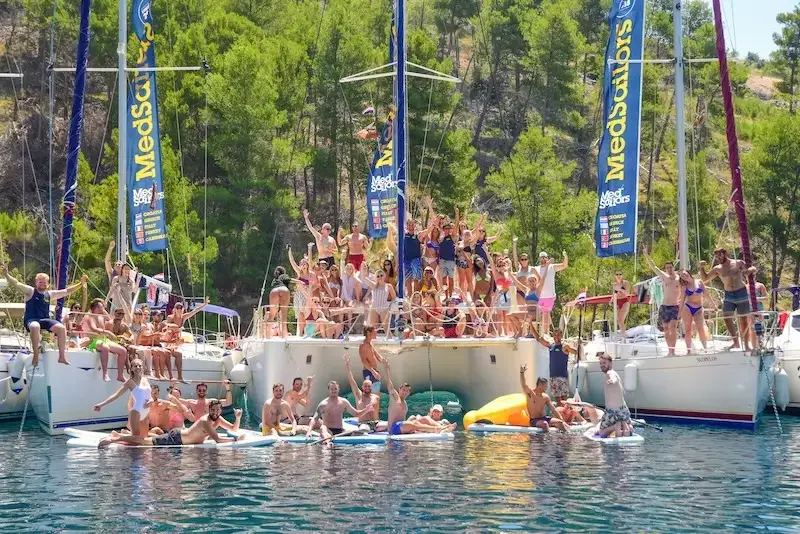Large group of MedSailors guests partying on yachts and inflatables in a secluded bay, surrounded by pine-covered hills during a Croatia island-hopping sailing holiday.