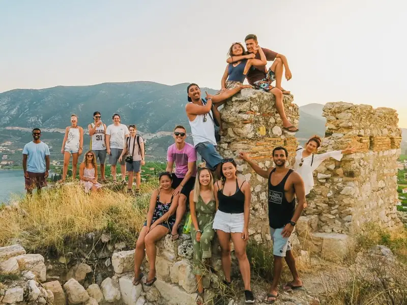 Group of young travellers posing on old stone ruins at sunset with mountains and a wide valley in the background, capturing the Peloponnese adventure travel vibe. Image used in a MedSailors Odyssey locations in Greece guide to support the Sparta and mainland Greece section, linking The Odyssey and the Trojan War world to real places you can visit today, including Sparta and nearby historic sites in the Peloponnese.