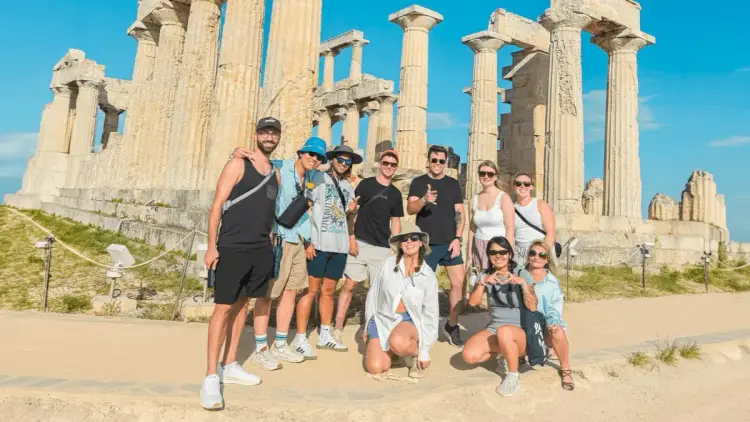 Group of MedSailors travellers smiling together in front of an ancient Greek temple on Aegina island, enjoying a cultural excursion during their Greece island hopping sailing holiday.